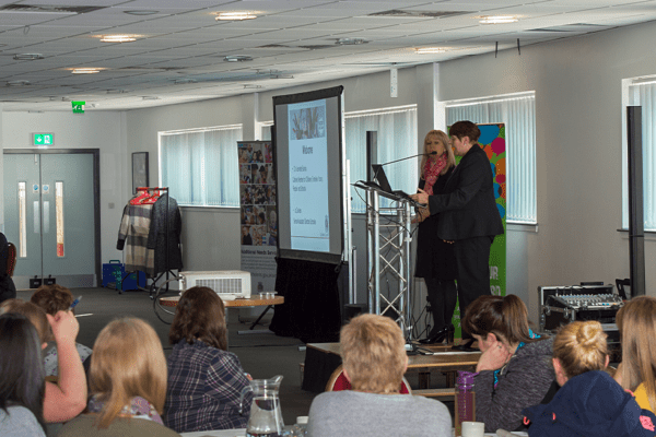 2 women speaking at an event Infront of people