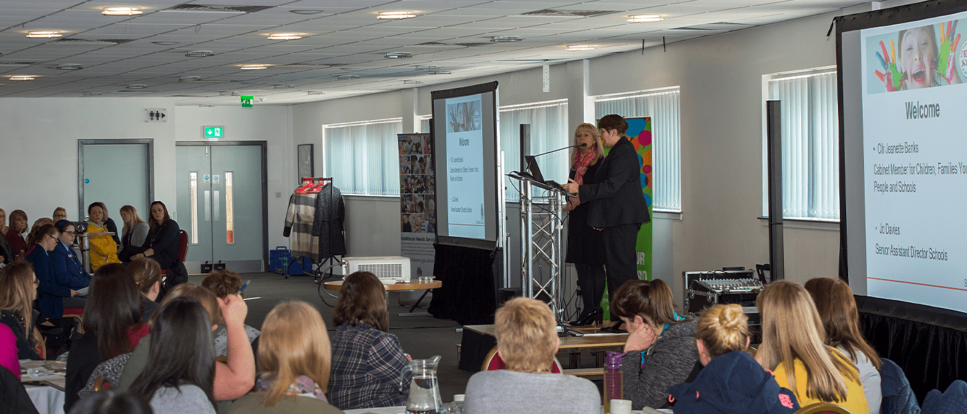 2 women speaking at an event Infront of people